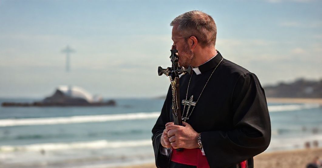 Catholic priest in traditional cassock at Bondi Beach after terrorist attack, holding crucifix with solemn expression.
