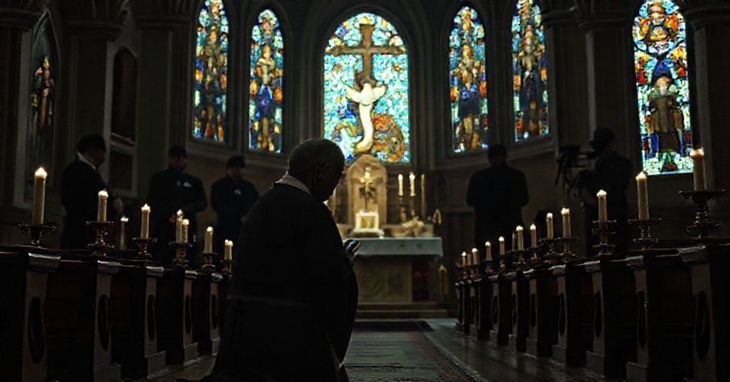 A traditional Catholic priest kneeling in prayer before a high altar in a dimly lit church as FBI agents monitor him in the background.